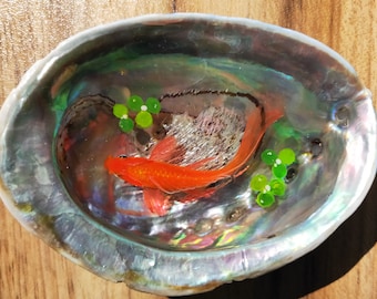 May include: A small orange fish swims in a clear resin shell with green beads. The shell has an iridescent finish.