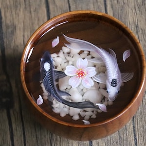 May include: A wooden bowl filled with water, white pebbles, and two resin fish. One fish is black with a white spot, and the other is white with a black spot. A pink cherry blossom flower is in the center of the bowl.
