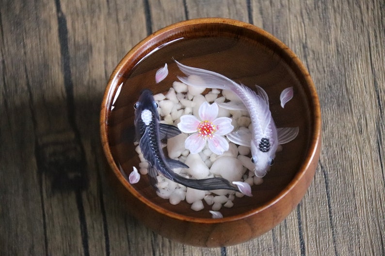 May include: A wooden bowl filled with water, white pebbles, and two resin fish. One fish is black with a white spot, and the other is white with a black spot. A pink cherry blossom flower is in the center of the bowl.
