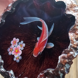 May include: A red and white goldfish swims in a resin art piece with a dark brown background. The resin art piece is shaped like a half circle and has a rough, textured edge. There are pink and white flowers painted on the resin.