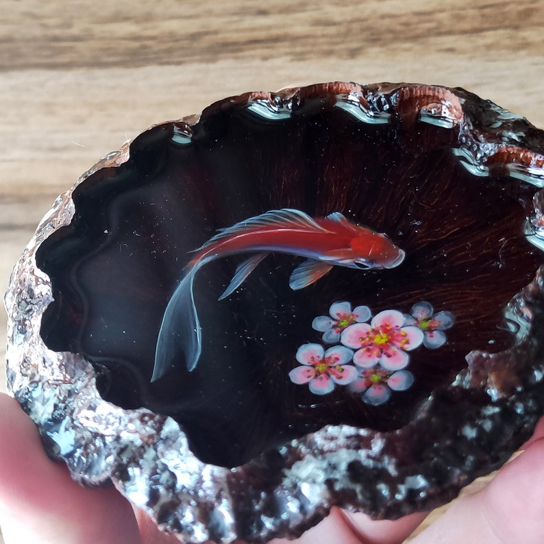 May include: A resin art piece featuring a red and white goldfish swimming in a dark blue pond with pink flowers. The pond is set in a dark brown, textured, circular frame.