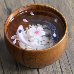 May include: A wooden bowl filled with water, white pebbles, and two resin koi fish. One fish is black with white spots, and the other is white with black spots. A pink cherry blossom is floating in the water.