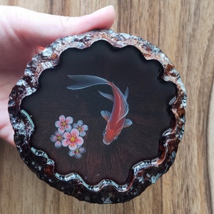 May include: A round wooden coaster with a black resin inlay featuring a red and white fish and pink flowers. The coaster has a wavy edge.