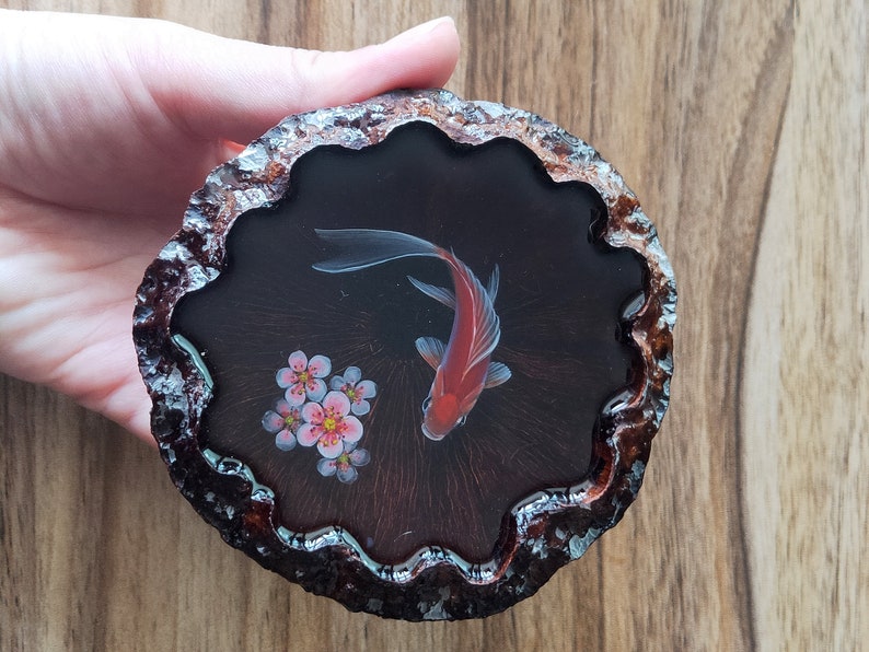 May include: A round wooden coaster with a black resin inlay featuring a red and white fish and pink flowers. The coaster has a wavy edge.