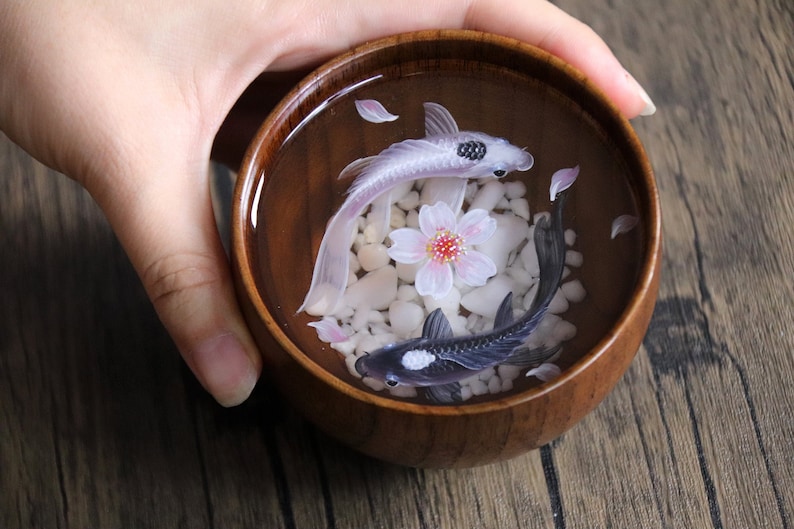 May include: A wooden bowl filled with water, white pebbles, and two resin fish. One fish is white with black accents, and the other is black with white accents. A pink cherry blossom is in the center of the bowl.