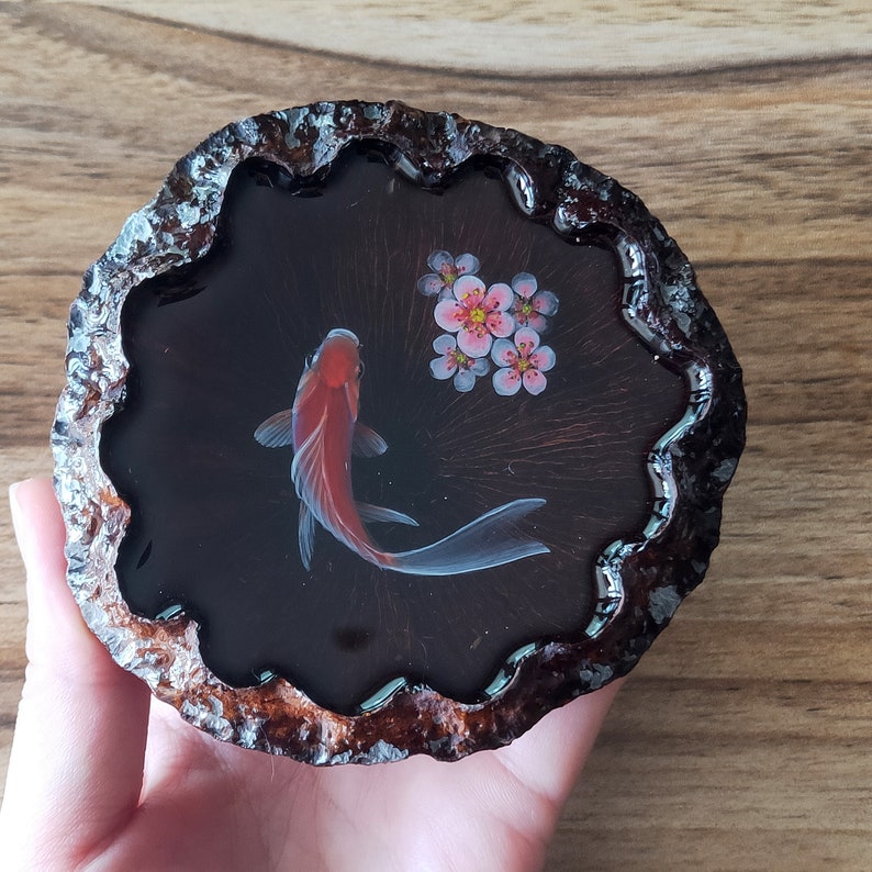 May include: A round wooden coaster with a dark brown resin inlay featuring a red and white goldfish and pink flowers.