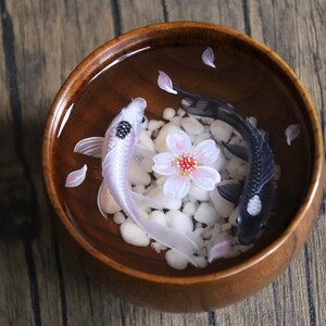 May include: A wooden bowl filled with water, white stones, and pink flower petals. Two koi fish, one black and one white, swim in the bowl.