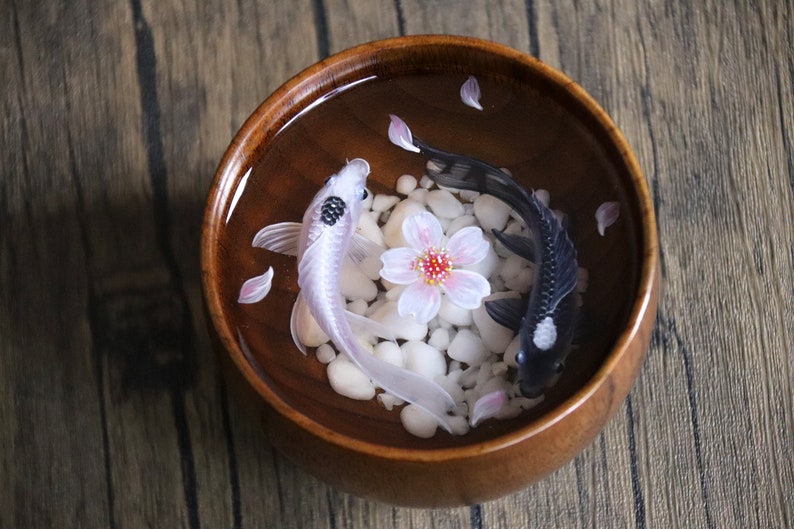 May include: A wooden bowl filled with water, white stones, and pink flower petals. Two koi fish, one black and one white, swim in the bowl.