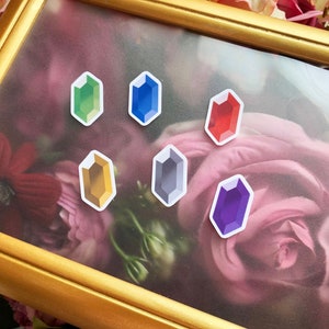 Peut inclure: Une affiche encadrée de six autocollants de gemmes colorées, chacun en forme d'hexagone allongé pointu. Les gemmes sont vertes, bleues, rouges, dorées, grises et violettes. Le cadre est doré et l'arrière-plan est une fleur rose.