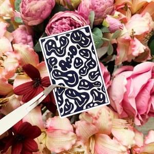 May include: A rectangular sticker featuring a pattern of navy blue smiley faces on a white background. The sticker is held by tweezers and is surrounded by pink, red, and white flowers.