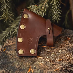 May include: A dark brown leather axe sheath featuring five brass-colored metal rivets along the curved edge, a secure strap with a matching metal stud closure, and a small embossed deer head logo on the side of the leather cover.