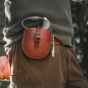 Könnte beinhalten: Eine braune Werkzeugtasche aus Leder mit Schnallenverschluss, befestigt an einem braunen Gürtel. Die Tasche enthält ein Gartengerät mit grünem Griff. Die Person trägt einen grünen Pullover und eine braune Hose.