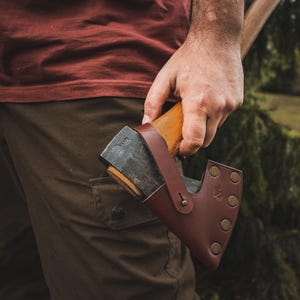 Può includere: Un'ascia con manico in legno e lama in metallo scuro, assicurata in un fodero di cuoio marrone. L'ascia è tenuta da una persona che indossa una camicia bordeaux e pantaloni verde oliva. La testa dell'ascia è contrassegnata dalle lettere "NB".