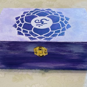 May include: A rectangular wooden box painted in shades of purple with a distressed finish. The lid features a white lotus flower design with a central Om symbol. A gold-colored metal latch is centered on the front of the box for closure.