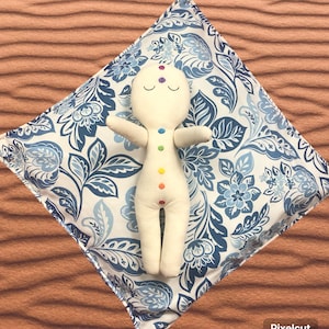 May include: A cream-colored doll with rainbow chakra dots and closed eyes rests on a blue and white floral patterned pillow. The pillow is square-shaped and features a detailed botanical design. The doll's arms are outstretched.