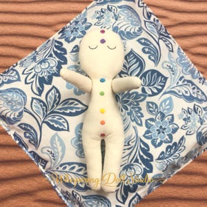 May include: A cream-colored fabric doll featuring seven small, circular, multi-colored embroidered chakra symbols along its center, resting on a square pillow with a blue and white floral pattern and the text Whispering Doll Studio.