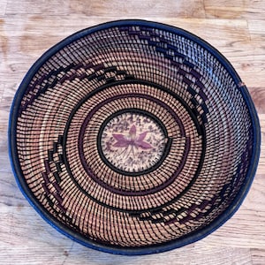May include: A round woven basket with a spiral design. The basket features a dark blue rim, black and purple accents, and a central floral design. The basket is made of natural fibers and sits on a wooden surface.