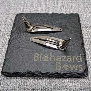 Op de afbeelding: Twee zilveren haarclips met puntige metalen spikes. De clips liggen op een zwart leisteen oppervlak met de tekst "Biohazard Bows" erop gegraveerd.