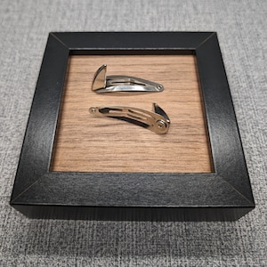May include: Two silver hair clips in a black frame with a wood background. The clips are both in the shape of a half-circle with a small, pointed end.