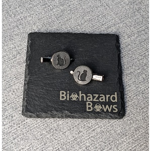 May include: Two cat silhouette cufflinks on a black slate coaster. The coaster is engraved with the words "Biohazard Bows" and a biohazard symbol. The cufflinks have a silver-tone finish.