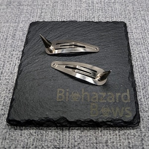 May include: Two silver hair clips with pointed ends. The clips are on a black slate surface with the text "Biohazard Bows" printed on it.