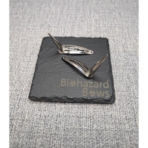 May include: Two silver hair clips with pointed ends, on a black slate surface with the text "Biohazard Bows" engraved in white.