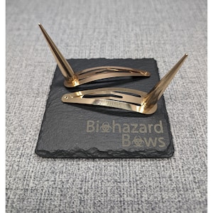 May include: Two gold hair clips with pointed ends. The clips are on a black slate surface with the text "Biohazard Bows" engraved on it.