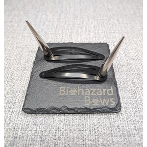 May include: Two black hair clips with silver pointed tips. The clips are on a black slate surface with the text "Biohazard Bows" engraved on it.