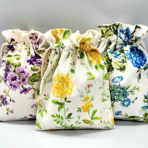 May include: Three drawstring bags with floral patterns. The bags are made of white fabric with different colored flowers. The first bag has purple flowers, the second has yellow flowers, and the third has blue flowers.