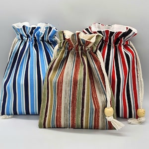 May include: Three drawstring bags with vertical striped patterns. One bag has blue and white stripes, another has a mix of brown, green, and red stripes, and the third has red, black, and white stripes. Each bag has a white drawstring with a wooden bead.