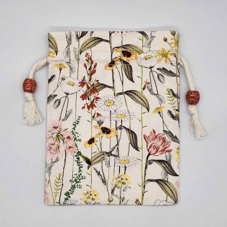 May include: A white drawstring bag with a floral print featuring red, yellow, and pink flowers. The bag has a white drawstring with brown wooden beads.