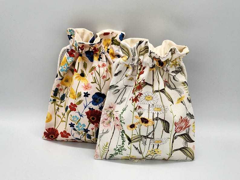 May include: Two drawstring bags with floral patterns. The bag on the left has a red, yellow, blue, and white floral pattern. The bag on the right has a white, yellow, and green floral pattern.