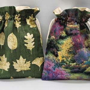 May include: Two drawstring bags. One is dark green with a pattern of light tan leaves. The other bag features a colorful landscape design with trees and foliage in shades of purple, yellow, and green. Both bags have cream-colored drawstrings.