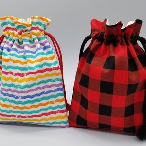 May include: Two drawstring bags. One bag features a white background with colorful horizontal wavy stripes in shades of blue, purple, yellow, orange, and pink. The other bag has a red and black buffalo plaid pattern.