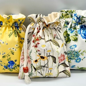 May include: Three drawstring fabric bags with floral patterns. The bags are made of white fabric with different floral prints. The first bag has a yellow background with blue and white flowers. The second bag has a white background with red, yellow, and white flowers. The third bag has a white background with blue and green flowers.