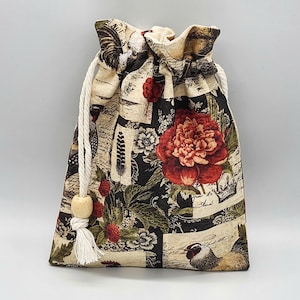 May include: A drawstring bag with a floral and botanical print. The bag features a large red peony, strawberries, and rooster illustrations on a black and cream background. A white drawstring with a wooden bead closure secures the top.