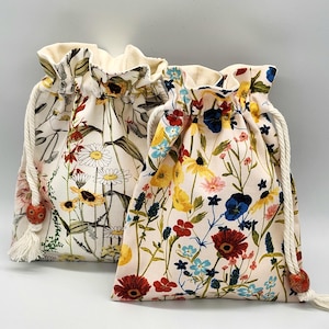 May include: Two drawstring bags with floral patterns. The bag on the left has a white background with brown, yellow, red, and white flowers. The bag on the right has a light pink background with red, yellow, blue, and white flowers.