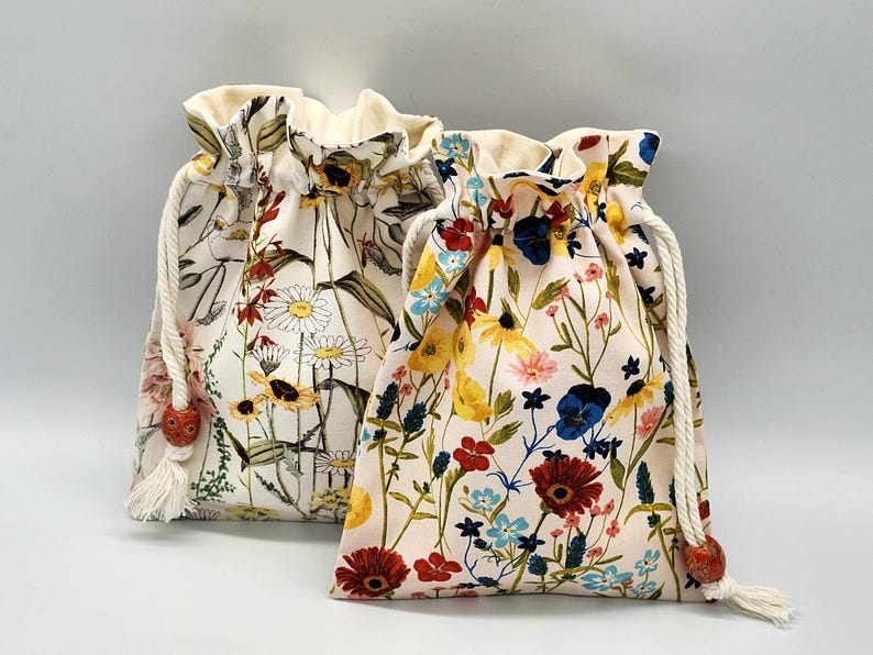 May include: Two drawstring bags with floral patterns. The bag on the left has a white background with brown, yellow, red, and white flowers. The bag on the right has a light pink background with red, yellow, blue, and white flowers.