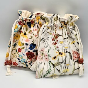 May include: Two drawstring bags with floral patterns. The bag on the left has a pink background with red, yellow, blue, and green flowers. The bag on the right has a white background with red, yellow, and green flowers.