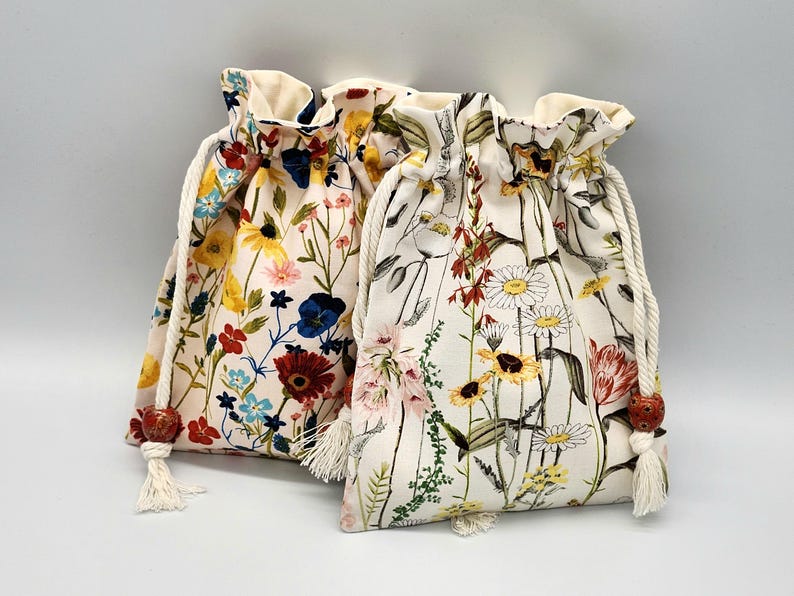 May include: Two drawstring bags with floral patterns. The bag on the left has a pink background with red, yellow, blue, and green flowers. The bag on the right has a white background with red, yellow, and green flowers.