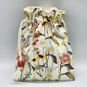 May include: A white drawstring bag with a floral print featuring various flowers, including daisies, tulips, and other wildflowers. The bag has a white tassel and a red bead.