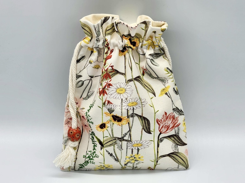 May include: A white drawstring bag with a floral print featuring various flowers, including daisies, tulips, and other wildflowers. The bag has a white tassel and a red bead.