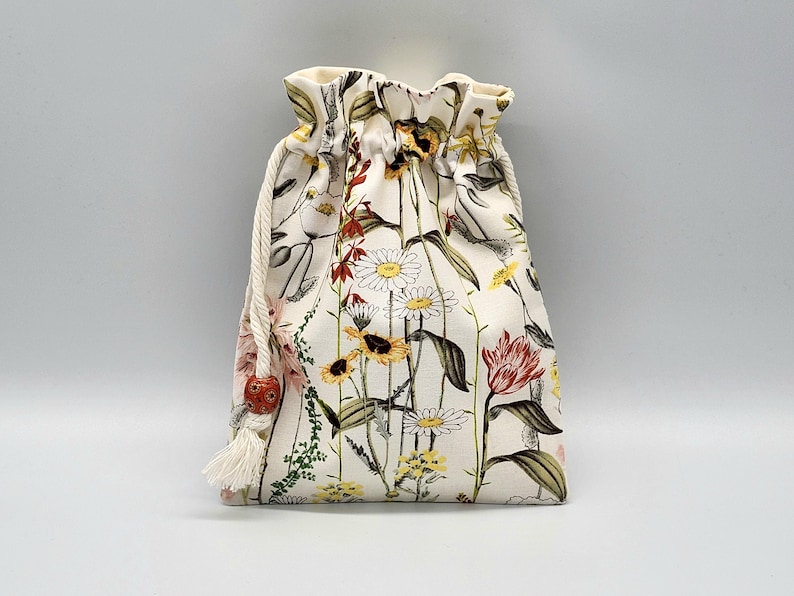 May include: A white drawstring bag with a floral print featuring various flowers, including daisies, sunflowers, and red tulips. The bag has a white drawstring with a red bead.