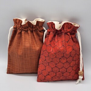 May include: Two drawstring bags made of fabric with different patterns. The bag on the left has a brown and orange geometric pattern. The bag on the right has a red and orange honeycomb pattern.