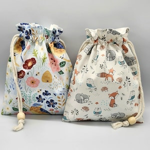 May include: Two drawstring bags with floral and woodland animal patterns. One bag features a light blue background with flowers, bees, and birds. The other bag has a cream background with foxes, owls, and hedgehogs. Both bags have a beige drawstring.