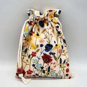 May include: A white drawstring bag with a floral print featuring red, yellow, blue, and pink flowers. The bag has a white cotton drawstring with a red tassel.
