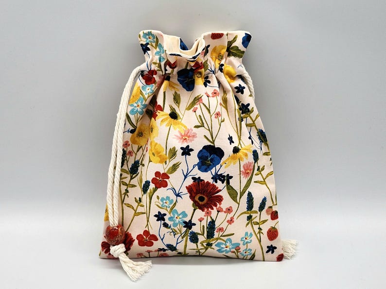 May include: A white drawstring bag with a floral print featuring red, yellow, blue, and pink flowers. The bag has a white cotton drawstring with a red tassel.