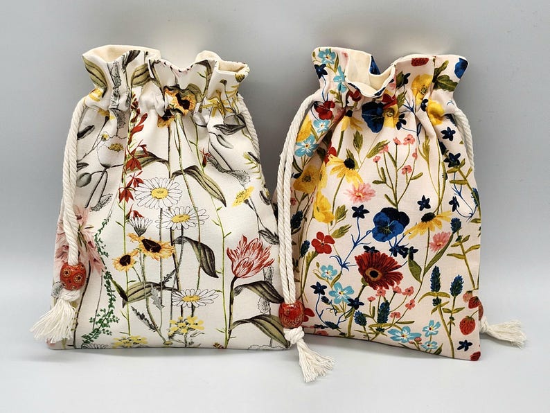 May include: Two white drawstring bags with floral patterns. The bag on the left has a red, yellow, and white floral pattern. The bag on the right has a blue, red, yellow, and white floral pattern.