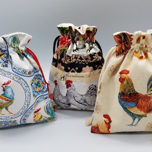 May include: Three drawstring bags with rooster designs. The first bag is white with blue and red rooster designs. The second bag is black and white with a rooster design and the text "Providence, RI". The third bag is beige with a red, orange, and blue rooster design.
