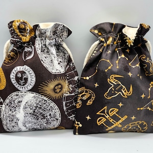 May include: Two drawstring bags with celestial and zodiac designs. One bag is black with gold constellations and zodiac signs, the other is brown with white constellations and sun faces.